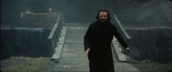 Movie still from “The Keep” (1983), directed by Michael Mann – A man in a black robe walking across a bridge; Medium shot, Over the shoulder angle