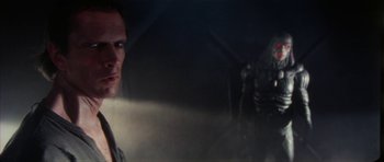 Movie still from “The Keep” (1983), directed by Michael Mann – A man standing next to another man in a dark room; Close Up shot, Over the shoulder angle