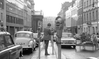 A black and white photo of a man standing on a sidewalk near parking meters