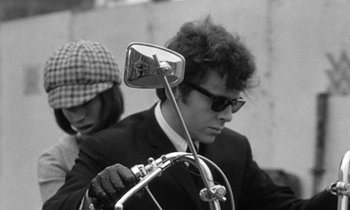 Movie still from “The Knack... and How to Get It” (1965), directed by Richard Lester – A man in a suit and sunglasses holding onto a motorcycle mirror; Medium shot, Low angle