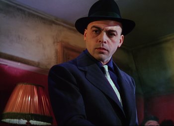 Movie still from “The Ladykillers” (1955), directed by Alexander Mackendrick – A man in a suit and hat standing in front of a lamp; Medium shot, Low angle
