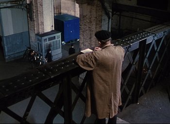 Movie still from “The Ladykillers” (1955), directed by Alexander Mackendrick – A man standing on the side of a bridge reading a book; Wide shot, High angle