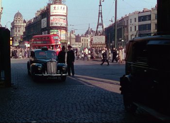 Movie still from “The Ladykillers” (1955), directed by Alexander Mackendrick – A car is driving down the street in a busy city; Extreme Wide shot, High angle