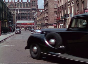 Movie still from “The Ladykillers” (1955), directed by Alexander Mackendrick – An old car driving down a street past tall buildings; Extreme Wide shot, High angle