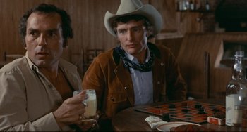 Movie still from “The Last Movie” (1971), directed by Dennis Hopper – A man wearing a cowboy hat sitting at a table with another man; Medium shot, Over the shoulder angle