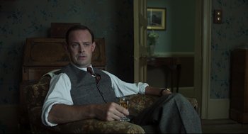 Movie still from “The Little Stranger” (2018), directed by Lenny Abrahamson – A man sitting in a chair holding a glass of wine; Medium shot, Low angle