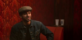 Movie still from “Always Be My Maybe” (2019), directed by Nahnatchka Khan – A man wearing a hat sitting on top of a red couch; Close Up shot, Over the shoulder angle