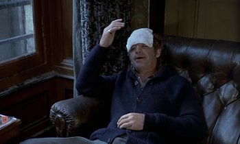 Movie still from “The Magnificent One” (1982), directed by Richard Greatrex – A man with a bandage on his head is sitting in a chair; Medium shot, High angle