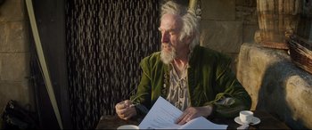 Movie still from “The Man Who Killed Don Quixote” (2018), directed by Terry Gilliam – An older man sitting at a table with a paper; Medium shot, Over the shoulder angle