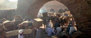 Movie still from “The Man Who Would Be King” (1975), directed by John Huston – A group of people standing around a building with a bunch of animals; Wide shot, High angle
