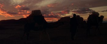 Movie still from “The Man Who Would Be King” (1975), directed by John Huston – A man standing next to a camel in the desert at sunset; Extreme Wide shot, Low angle