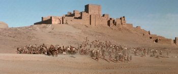 Movie still from “The Man Who Would Be King” (1975), directed by John Huston – A large group of people are gathered in the desert near a castle; Extreme Wide shot, High angle