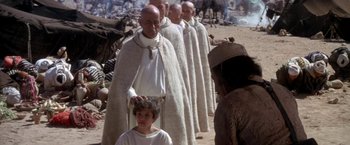 Movie still from “The Man Who Would Be King” (1975), directed by John Huston – A group of men standing next to each other on top of a dirt field; Medium shot, Over the shoulder angle