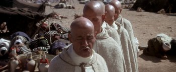 Movie still from “The Man Who Would Be King” (1975), directed by John Huston – A group of bald men standing next to each other on a beach; Close Up shot, High angle