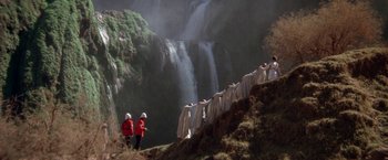 Movie still from “The Man Who Would Be King” (1975), directed by John Huston – A group of people standing next to a large waterfall; Extreme Wide shot, High angle