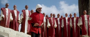 Movie still from “The Man Who Would Be King” (1975), directed by John Huston – A man in a red uniform stands in front of a group of men; Wide shot, Low angle