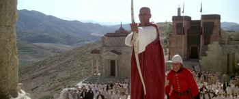 Movie still from “The Man Who Would Be King” (1975), directed by John Huston – A man holding a bow and arrow in front of a group of people; Medium shot, Low angle