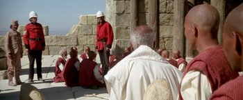 Movie still from “The Man Who Would Be King” (1975), directed by John Huston – A group of men sitting around a building; Wide shot, Over the shoulder angle