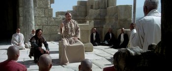 Movie still from “The Man Who Would Be King” (1975), directed by John Huston – A man sitting on top of a chair in front of a group of people; Wide shot, High angle