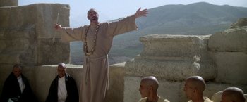 Movie still from “The Man Who Would Be King” (1975), directed by John Huston – An old man in a robe stands with his arms outstretched and a bald man sitting on the side of a building; Medium shot, Low angle