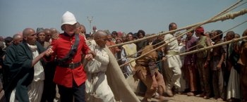 Movie still from “The Man Who Would Be King” (1975), directed by John Huston – A group of people in costume walking through a crowd of onlookers; Wide shot, Over the shoulder angle