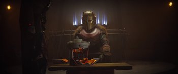 Movie still from “The Mandalorian” (2019), created by Jon Favreau – A man in a helmet sitting in front of a fire; Wide shot, Low angle