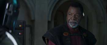 Movie still from “The Mandalorian” (2019), created by Jon Favreau – An older man wearing a red shirt and a black jacket; Close Up shot, Over the shoulder angle