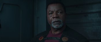 Movie still from “The Mandalorian” (2019), created by Jon Favreau – An older man wearing a red shirt and a black jacket; Close Up shot, Over the shoulder angle