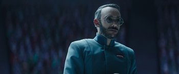 Movie still from “The Mandalorian” (2019), created by Jon Favreau – A man with a beard wearing a suit and round glasses; Close Up shot, Low angle