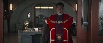 Movie still from “The Mandalorian” (2019), created by Jon Favreau – A man in a red and black outfit standing in a room; Medium shot, Over the shoulder angle