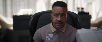 Movie still from “The Mandalorian” (2019), created by Jon Favreau – A man sitting in a chair with a button on his chest; Close Up shot, Over the shoulder angle