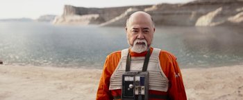 Movie still from “The Mandalorian” (2019), created by Jon Favreau – An older man wearing an orange shirt and holding a camera; Close Up shot, Over the shoulder angle