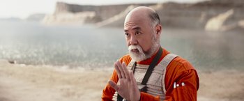 Movie still from “The Mandalorian” (2019), created by Jon Favreau – An older man wearing an orange shirt clapping his hands; Medium shot, Over the shoulder angle