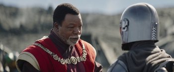 Movie still from “The Mandalorian” (2019), created by Jon Favreau – An older man wearing a red vest and a gold necklace; Close Up shot, Over the shoulder angle