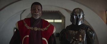 Movie still from “The Mandalorian” (2019), created by Jon Favreau – A man in a red and black outfit standing next to another man in a red and black outfit; Medium shot, Over the shoulder angle