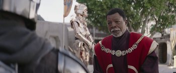 Movie still from “The Mandalorian” (2019), created by Jon Favreau – A man in a red and black outfit standing in front of a metal statue; Close Up shot, Over the shoulder angle