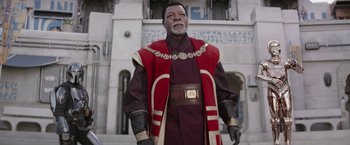 Movie still from “The Mandalorian” (2019), created by Jon Favreau – An older man in a red and black outfit stands in front of a white building; Medium shot, Low angle