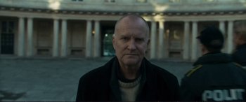 Movie still from “The Marco Effect” (2021), directed by Martin Zandvliet – A man with a bald head is standing in front of a building; Medium shot, Low angle