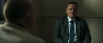 Movie still from “The Marco Effect” (2021), directed by Martin Zandvliet – A man wearing a suit and tie sitting in front of a wall; Medium shot, Over the shoulder angle