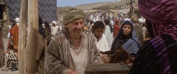 Movie still from “The Message” (1976), directed by Moustapha Akkad – An old man sitting at a table in a crowd of people; Medium shot, Over the shoulder angle