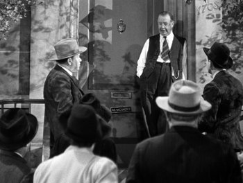 Movie still from “The More the Merrier” (1943), directed by George Stevens – Two men in suits and hats standing in front of a building; Medium shot, Over the shoulder angle
