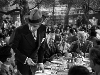 Movie still from “The More the Merrier” (1943), directed by George Stevens – A man in a hat standing at a table with a plate of food; Medium shot, Over the shoulder angle
