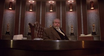 Movie still from “The Muppet Movie” (1979), directed by James Frawley – An older man sitting at a desk in a suit and tie; Wide shot, Low angle