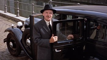 Movie still from “The Nun's Story” (1959), directed by Fred Zinnemann – An older man wearing a suit and a hat standing next to an old car; Medium shot, Low angle