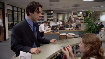 Movie still from “The Office” (2005), created by Ricky Gervais – A man in a suit and tie at a counter; Medium shot, Over the shoulder angle