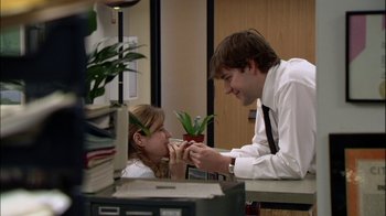 Movie still from “The Office” (2005), created by Ricky Gervais – A man and a woman sitting at a desk with a potted plant; Medium shot, High angle