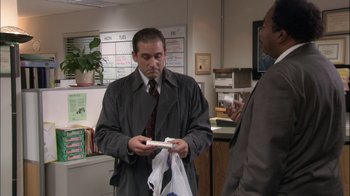 Movie still from “The Office” (2005), created by Ricky Gervais – A man in a suit and tie looking at an item in a bag; Medium shot, Over the shoulder angle