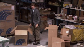 Movie still from “The Office” (2005), created by Ricky Gervais – A man in a suit and tie standing in a room with cardboard boxes; Wide shot, High angle