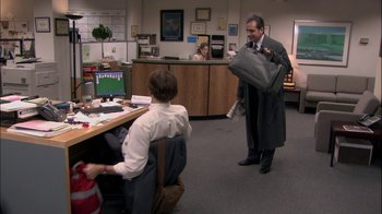 Movie still from “The Office” (2005), created by Ricky Gervais – A man is holding a bag in an office setting; Wide shot, High angle