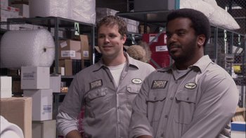 Movie still from “The Office” (2005), created by Ricky Gervais – A couple of men standing next to each other in a warehouse; Medium shot, Over the shoulder angle